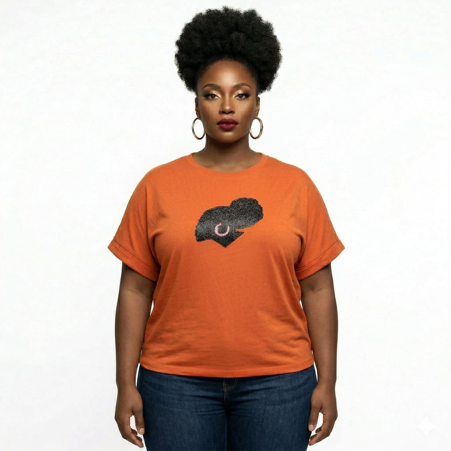 Woman in an orange t-shirt with a black rhinestone and metallic film design, dark jeans, earrings.