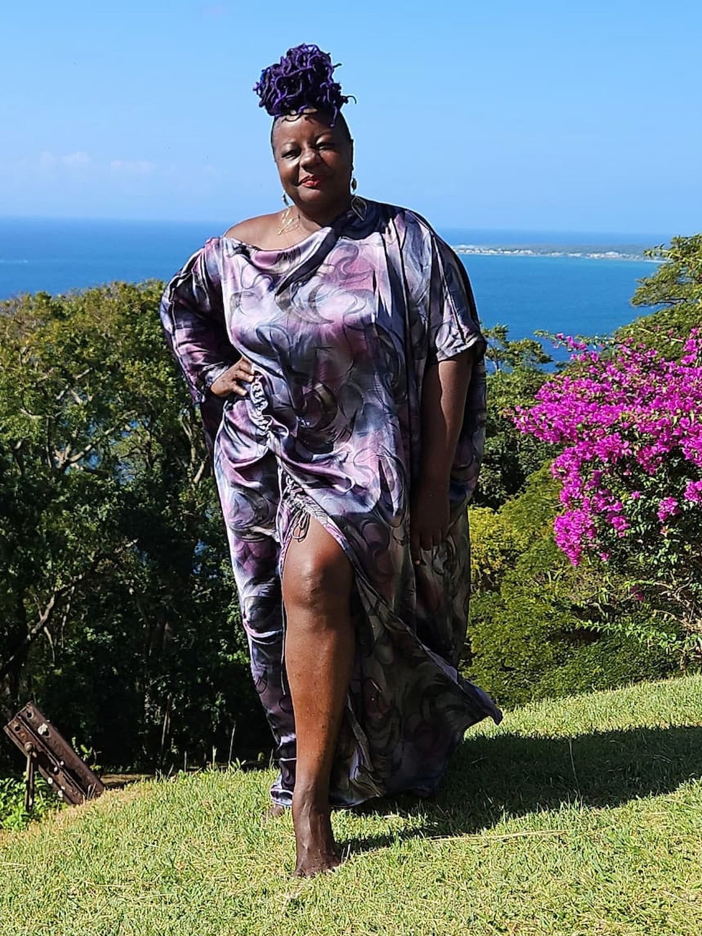 Woman in a patterned dress standing on a grassy hill with ocean and flowers in the background