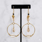 Pair of gold hoop earrings with shell accents on a black stand against a white background
