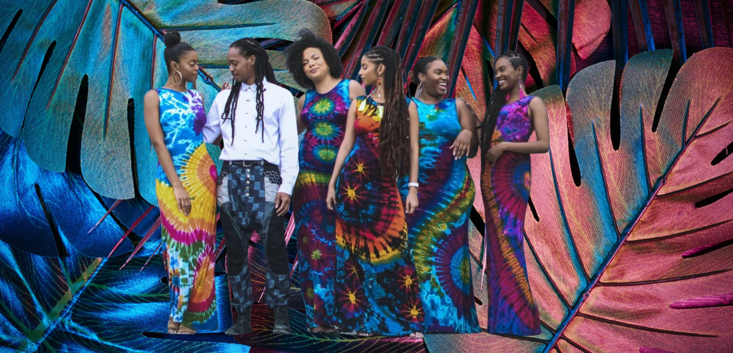 Group of people in colorful tie-dye outfits standing against a vibrant leaf-patterned background-Sanjules
