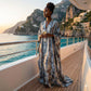 Woman in a long, silk satin kaftan standing on a yacht with a scenic coastal view.