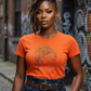 Woman in orange t-shirt with rhinestone loc design, dark wash jeans, and brown belt.