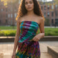 Woman wearing a tie-dye tube top and pants standing outdoors with buildings and greenery in the back