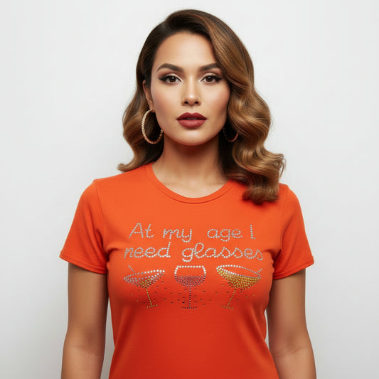 Orange women's combed cotton t-shirt with "At my age I need glasses" rhinestone design and cocktail glasses.
