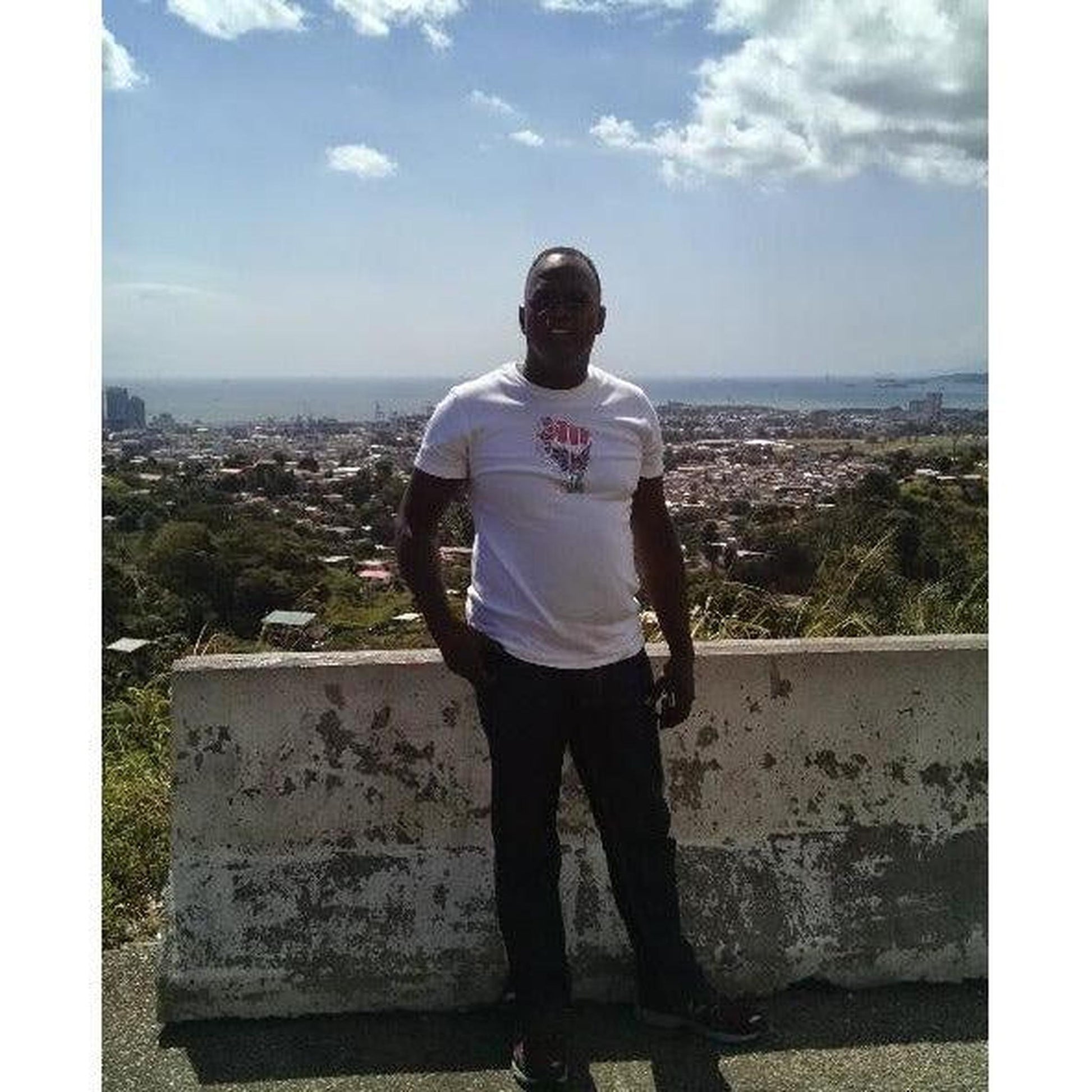 Man in white Black Power Fist Rhinestone T-Shirt, black pants, outdoors with city and ocean view.