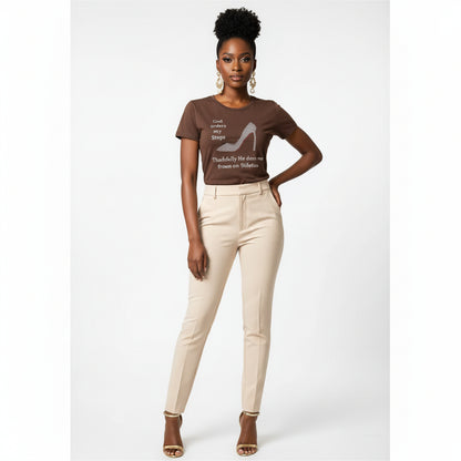 Woman in a brown "God Orders My Steps" rhinestone stiletto tee, beige pants, and gold heels.