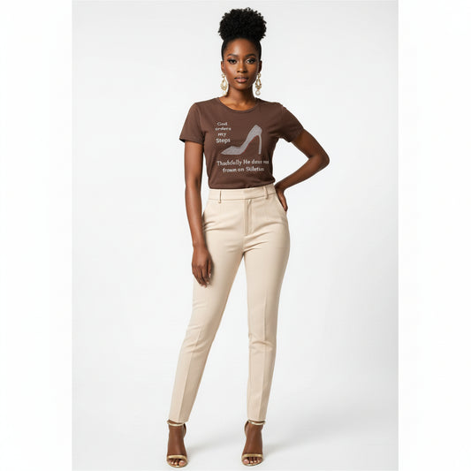 Woman in a brown "God Orders My Steps" rhinestone stiletto tee, beige pants, and gold heels.