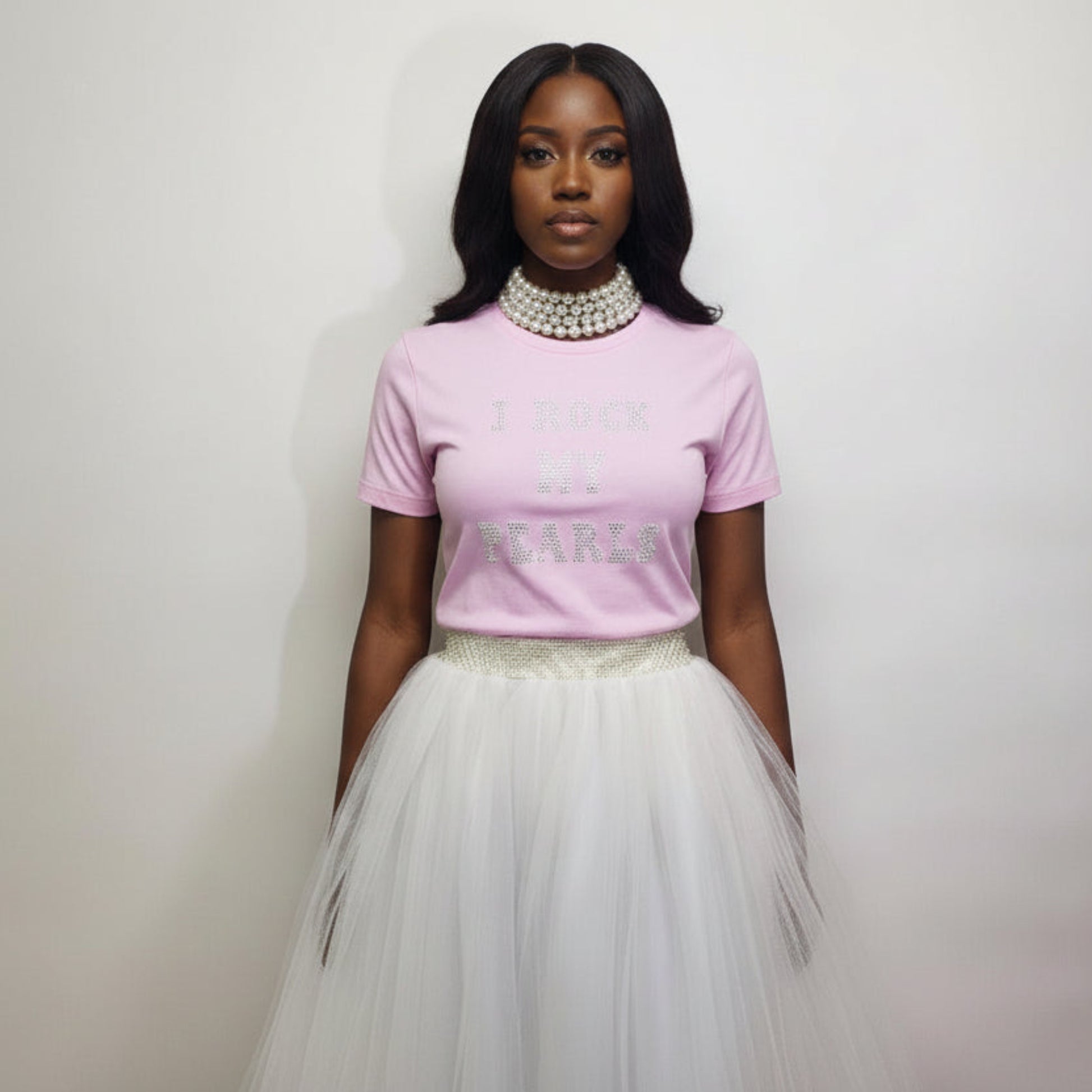 Woman wearing a pink t shirt with a rhinestone text and a white tulle skirt against a plain background